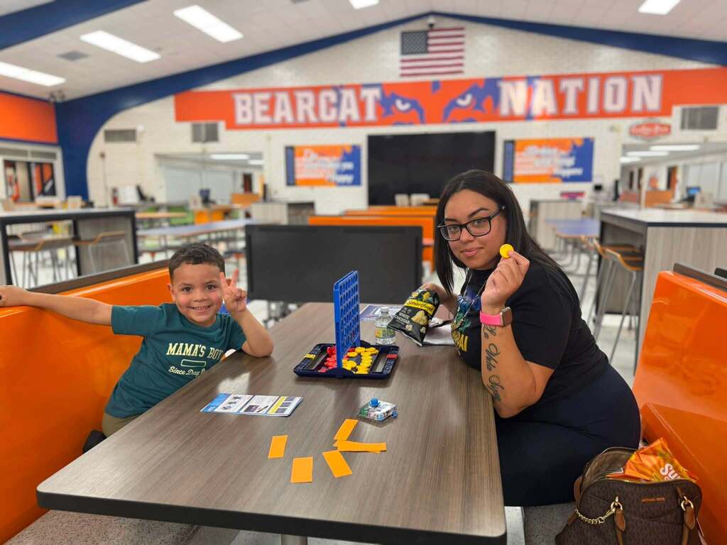 A student and an adult are playing Connect Four while sitting at a table in a school cafeteria.