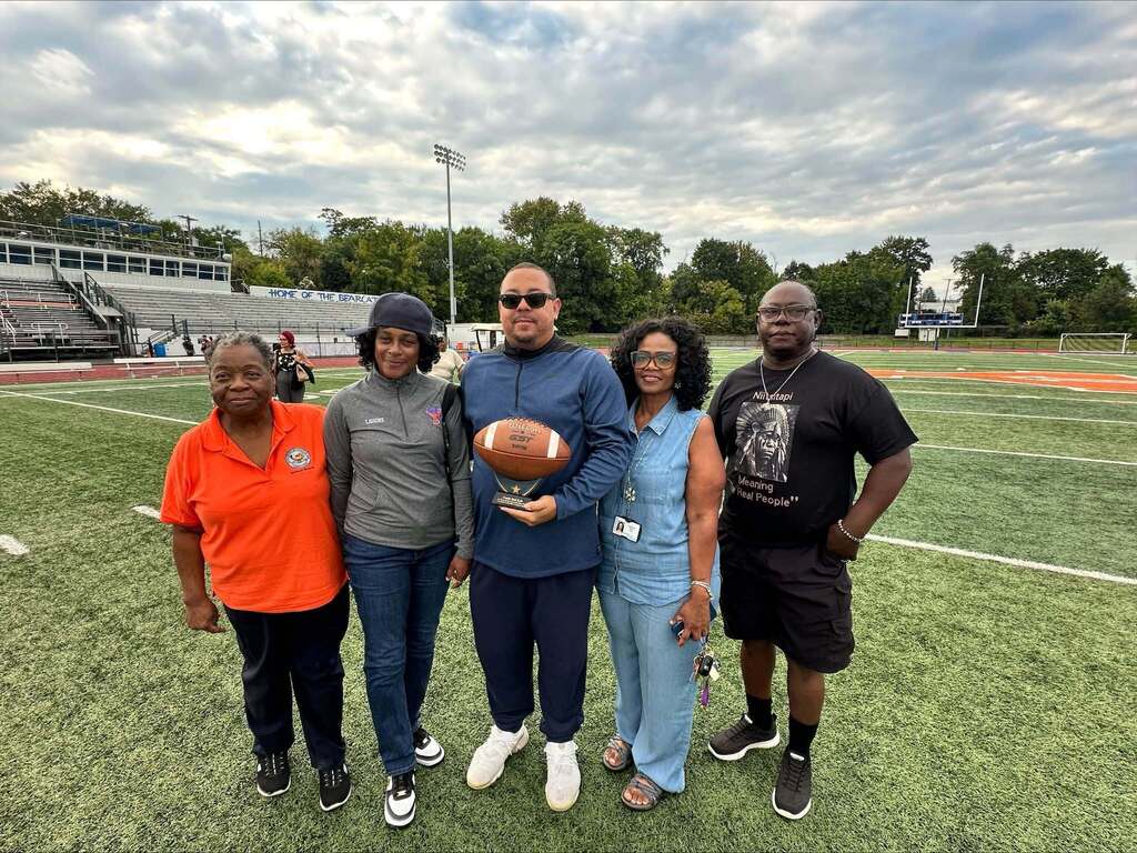 Five people, consisting of School Board Members and district staff members are posing together on a football field, with one district staff member holding a football.