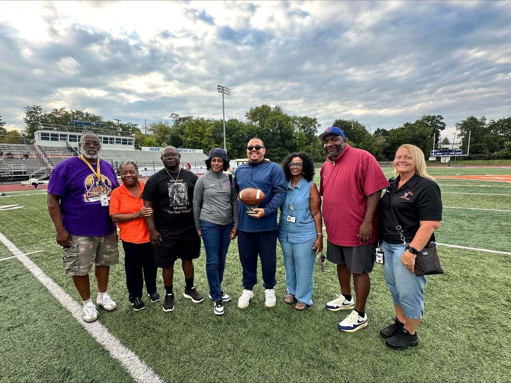 Eight people, consisting of School Board Members and district staff members are posing together on a football field, with one district staff member holding a football.