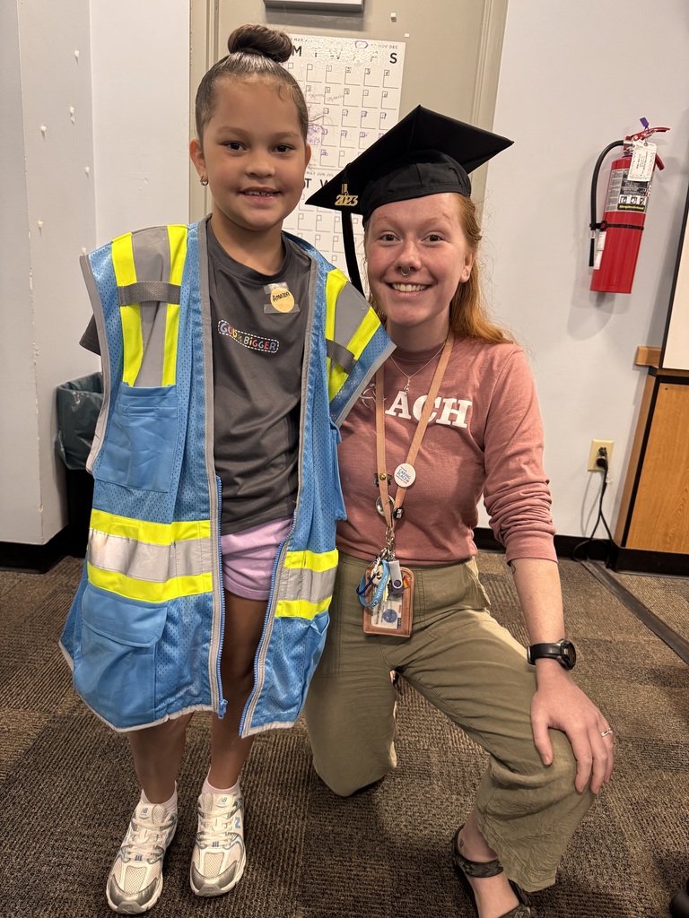 A young student in a safety vest is posing beside a district staff member wearing a graduation cap and gown.