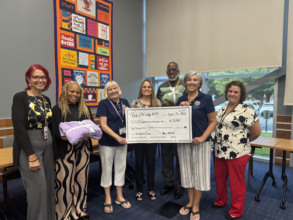 A group of six people, including district administrators, staff members, and local community members are posing together holding a blanket and a $2,000 donation check from the York Elks Lodge #213.