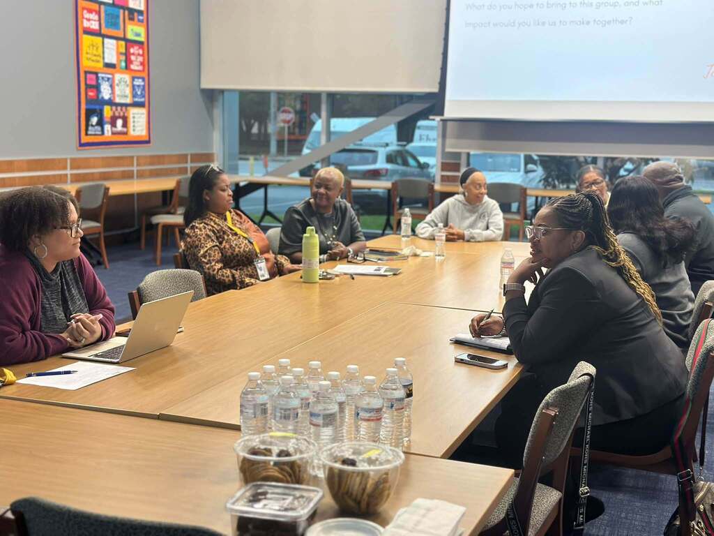 A group of district administrators, district staff, and community members are gathered around a table for a meeting, with laptops, notebooks, and refreshments.