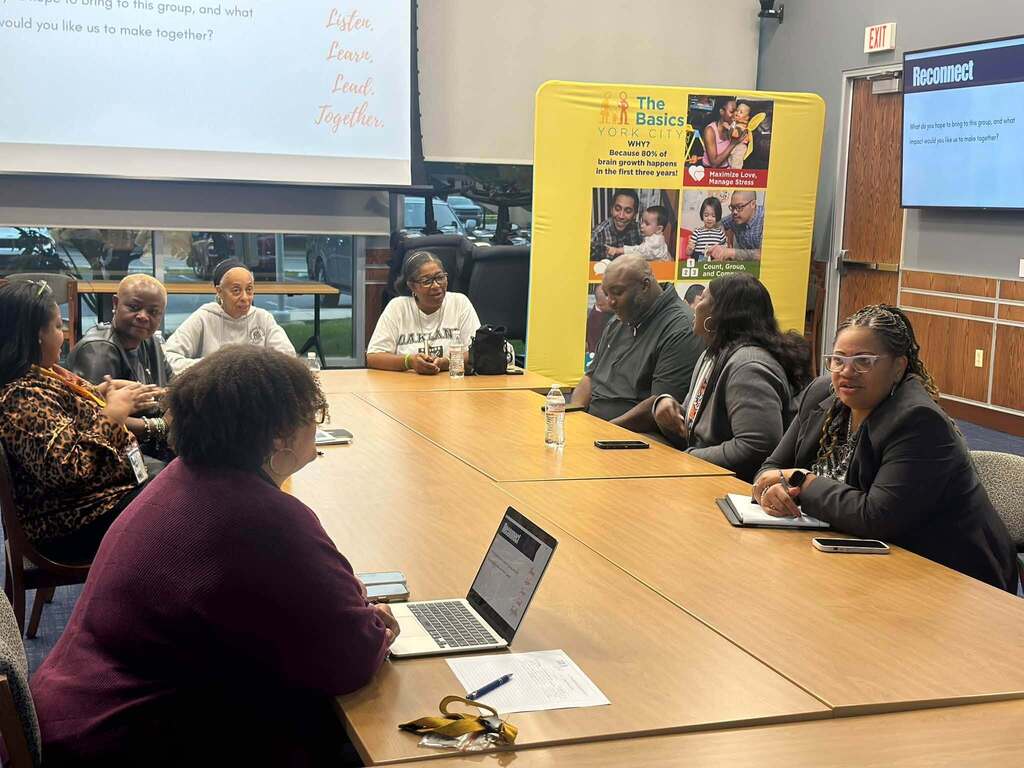 A group of district administrators, district staff, and community members are gathered around a table for a meeting, with laptops, notebooks, and refreshments.