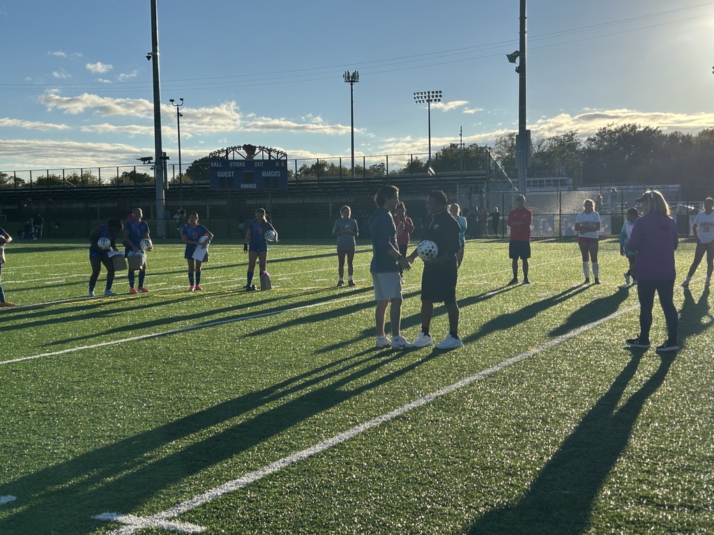 A group of soccer players and coaches are standing on an outdoor turf field.