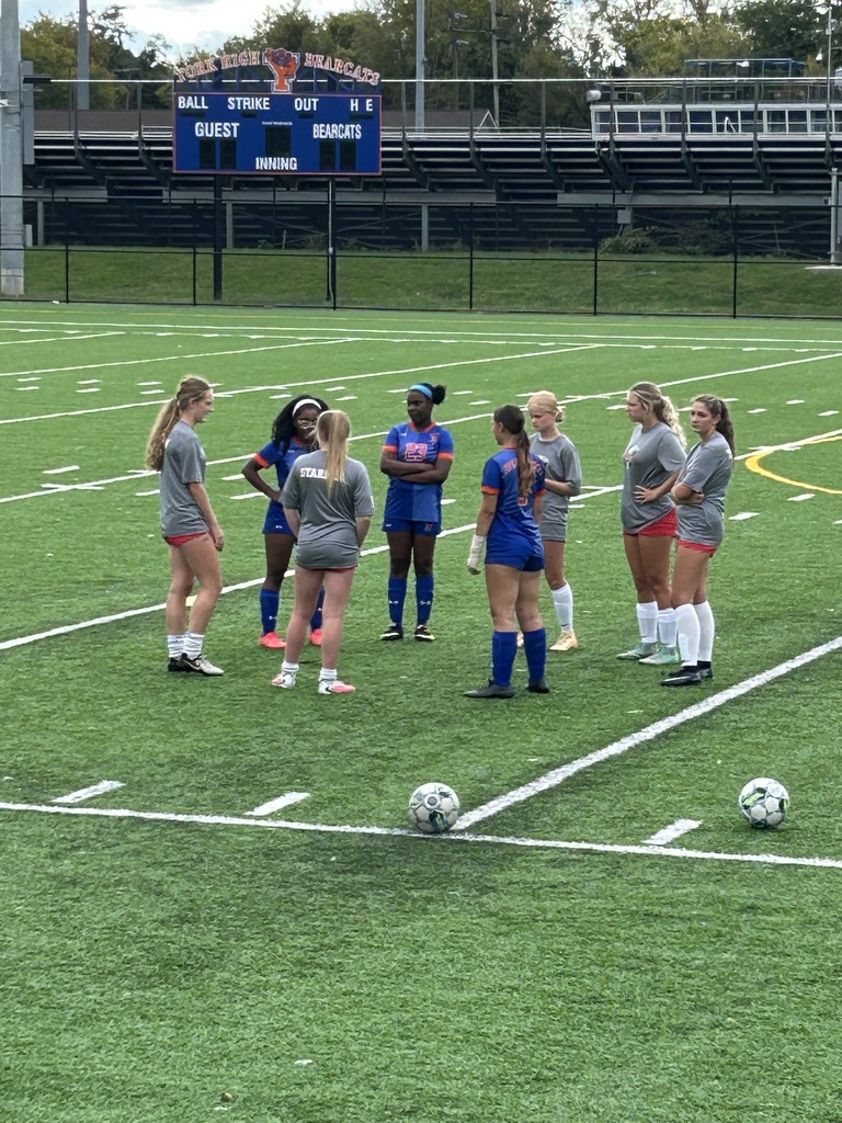 A group of soccer players are gathered on a turf field near the scoreboard.