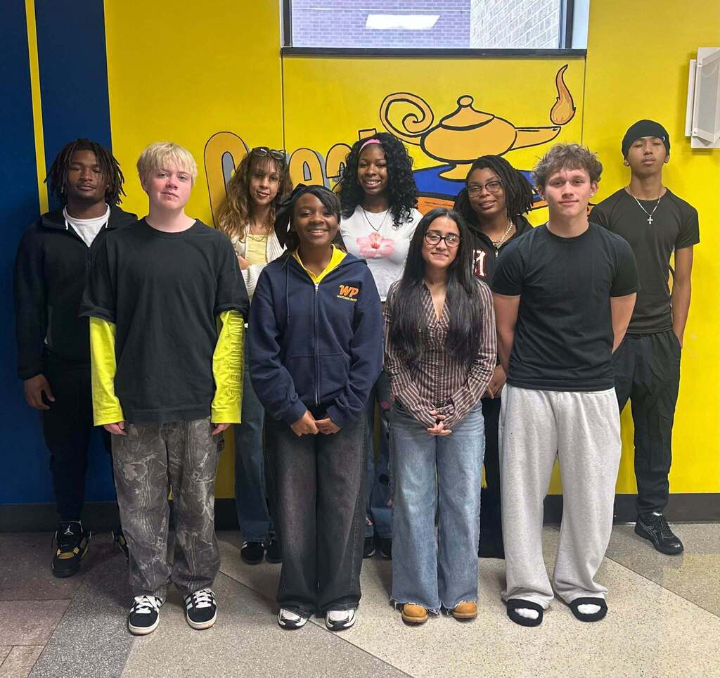 Eight students are posing for a group photo in front of a colorful school mural.