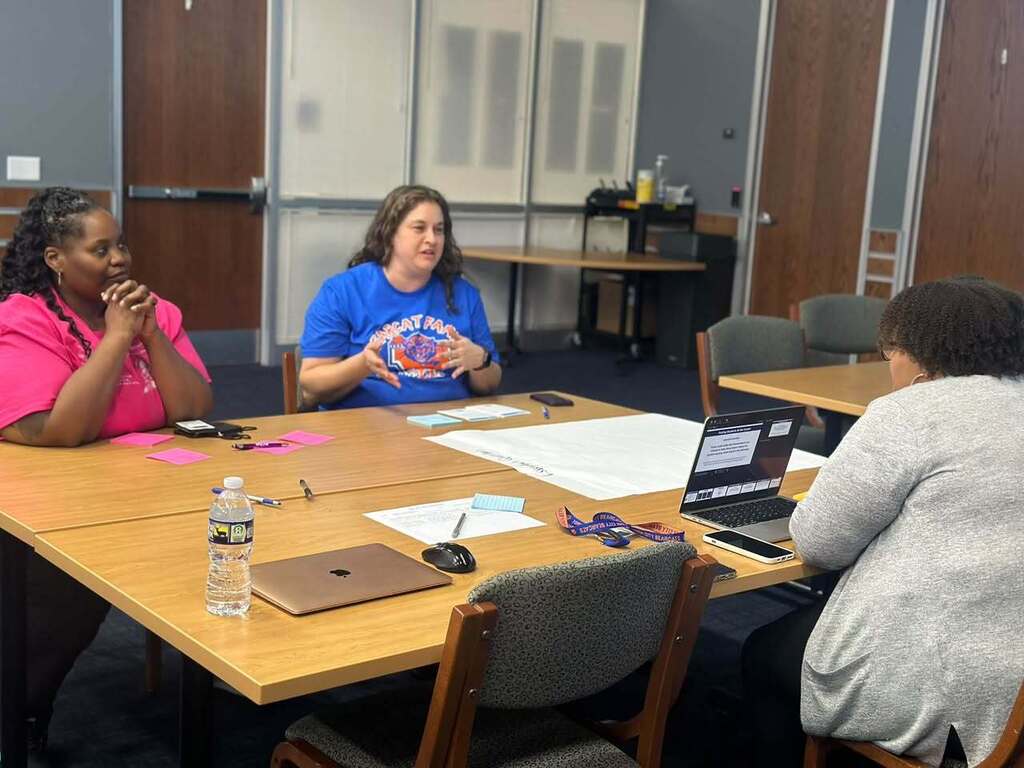 Three women are participating in a collaborative meeting around a table with laptops, notes, and other materials.