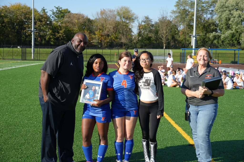 A William Penn girls’ soccer senior is posing with a family member, a teammate, and district staff members, while holding a framed photo during Senior Night.