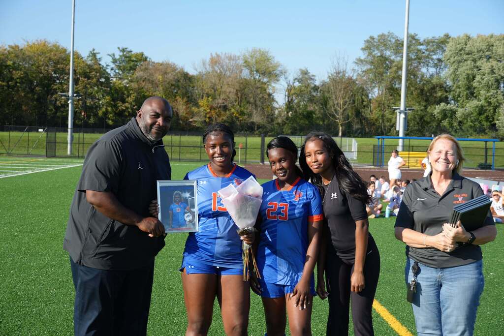 A William Penn girls’ soccer senior is posing with a family member, a teammate, and district staff members, while holding a framed photo during Senior Night.