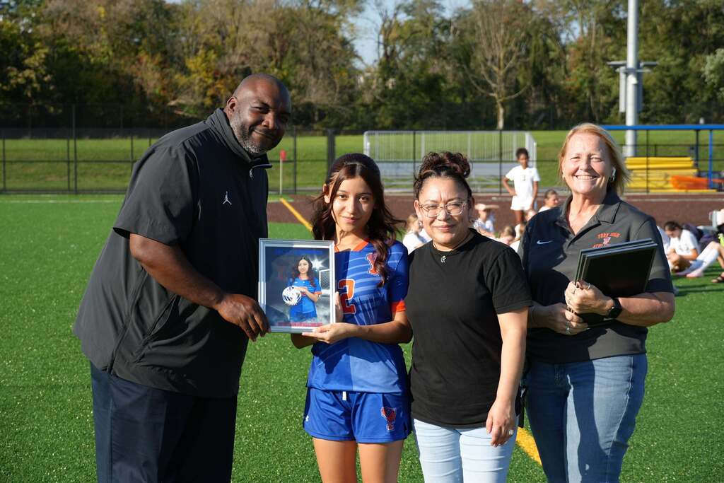A William Penn girls’ soccer senior is posing with a family member and district staff members, while holding a framed photo during Senior Night.
