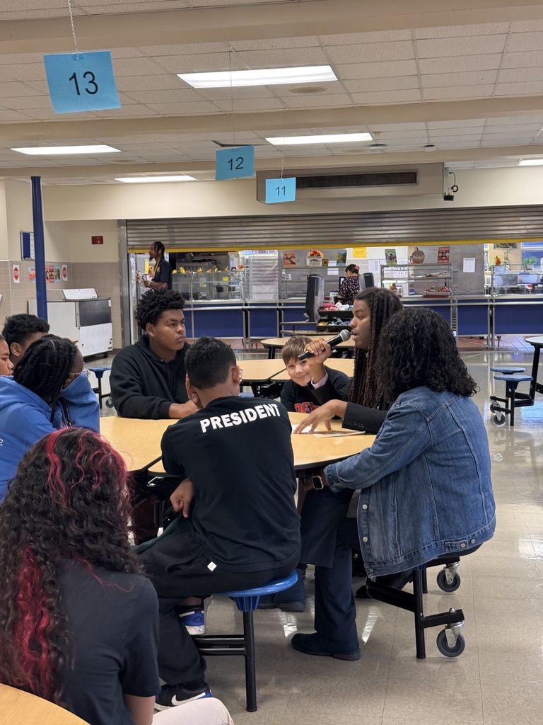 Students are gathered around a school cafeteria table listening to a guest speaker.