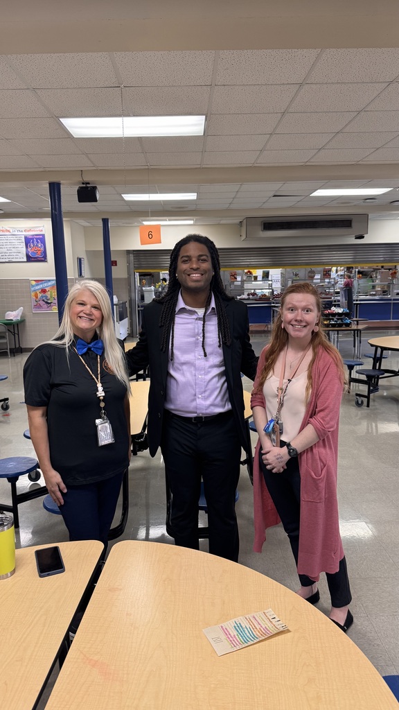 Two staff members and a guest speaker are smiling for a photo together in a school cafeteria.