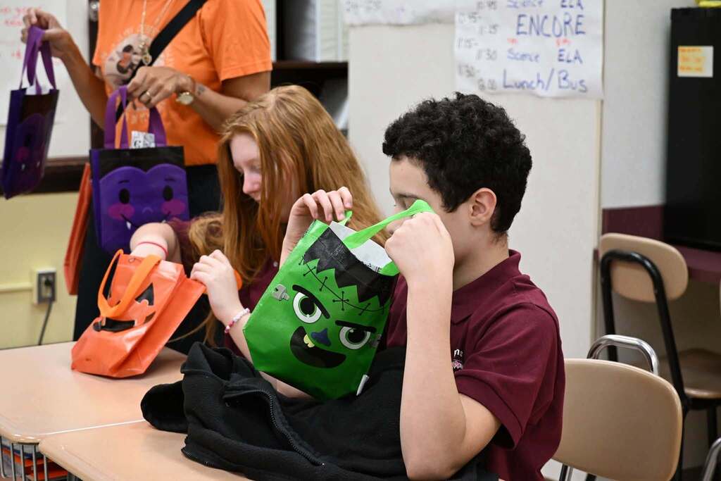 Two students are examining a green Frankenstein-themed gift bag and an orange pumpkin-themed gift bag while sitting at desks in a school classroom.