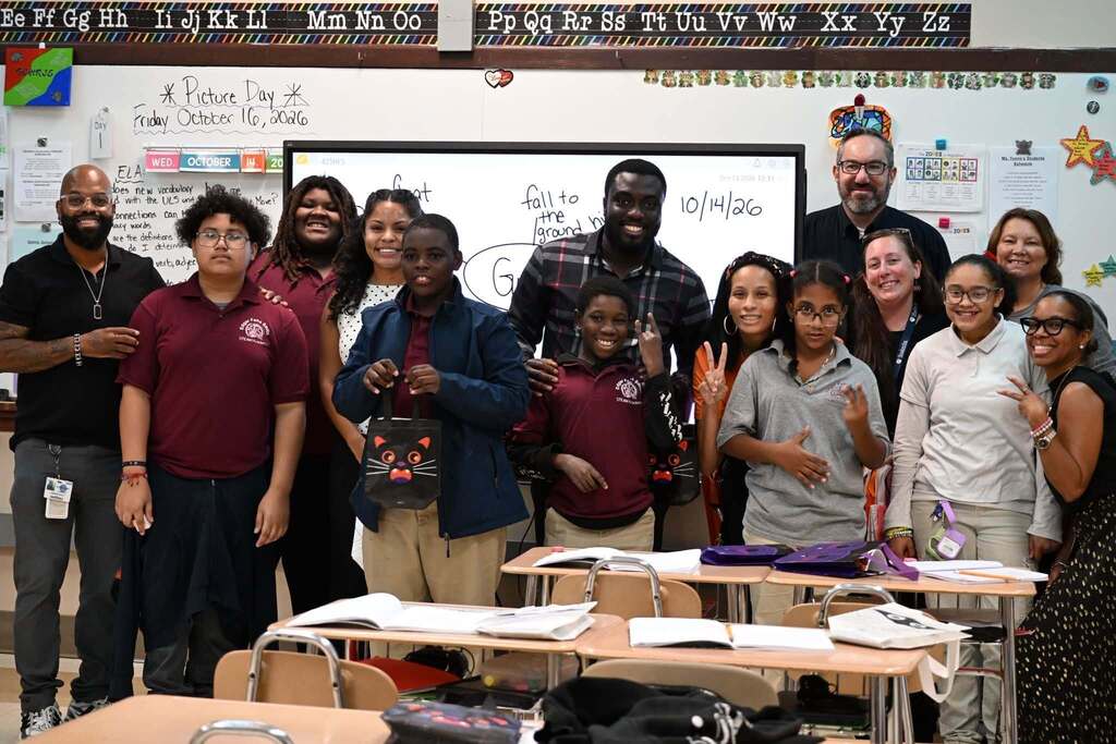 A group of district staff members, students, and community members are posing for a group photo in a school classroom, while smiling and holding colorful bags.