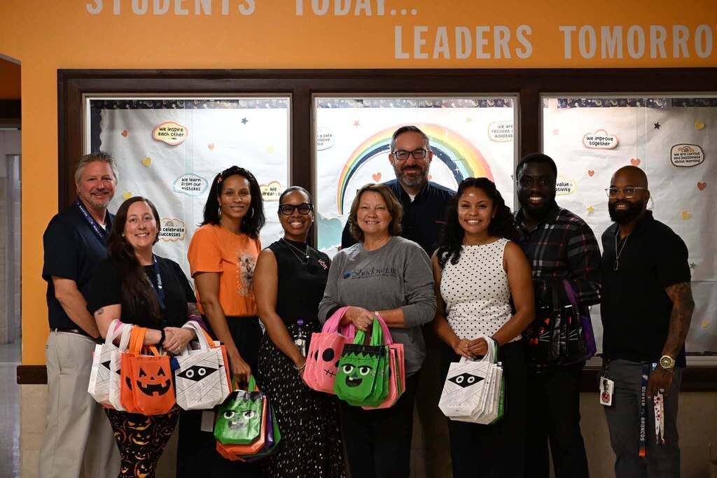 A group of district staff members and local community members are posing for a group photo while holding Halloween-themed treat bags and standing in front of a colorful display.