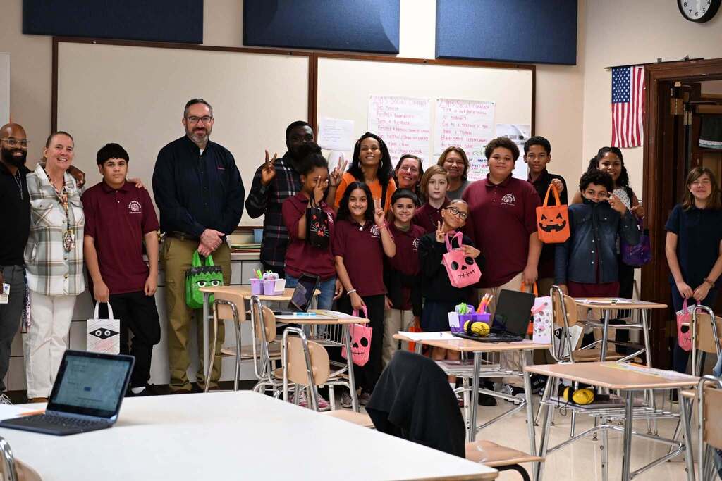 A group of district staff members, students, and community members are posing for a group photo in a school classroom, while smiling and holding colorful bags.