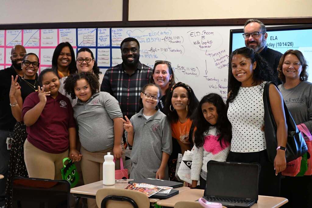 A group of district staff members, students, and community members are posing for a group photo in a school classroom, while smiling and holding colorful bags. Is this conversation helpful so far?