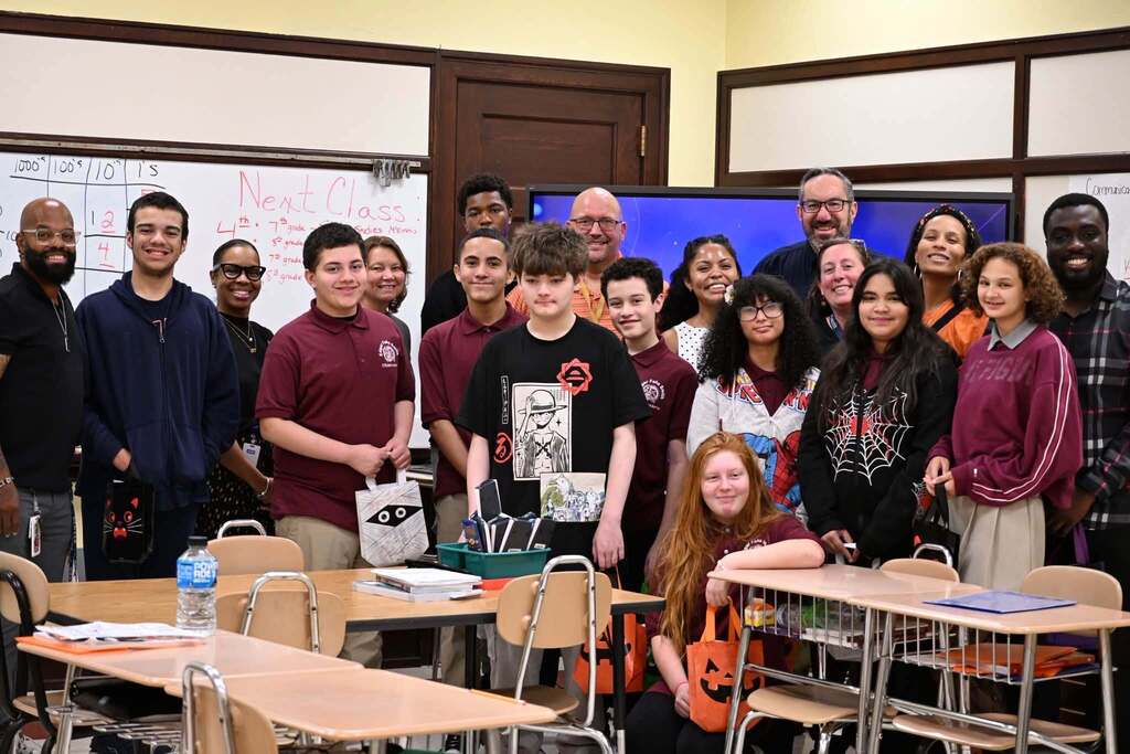 A group of district staff members, students, and community members are posing for a group photo in a school classroom, while smiling and holding colorful bags.