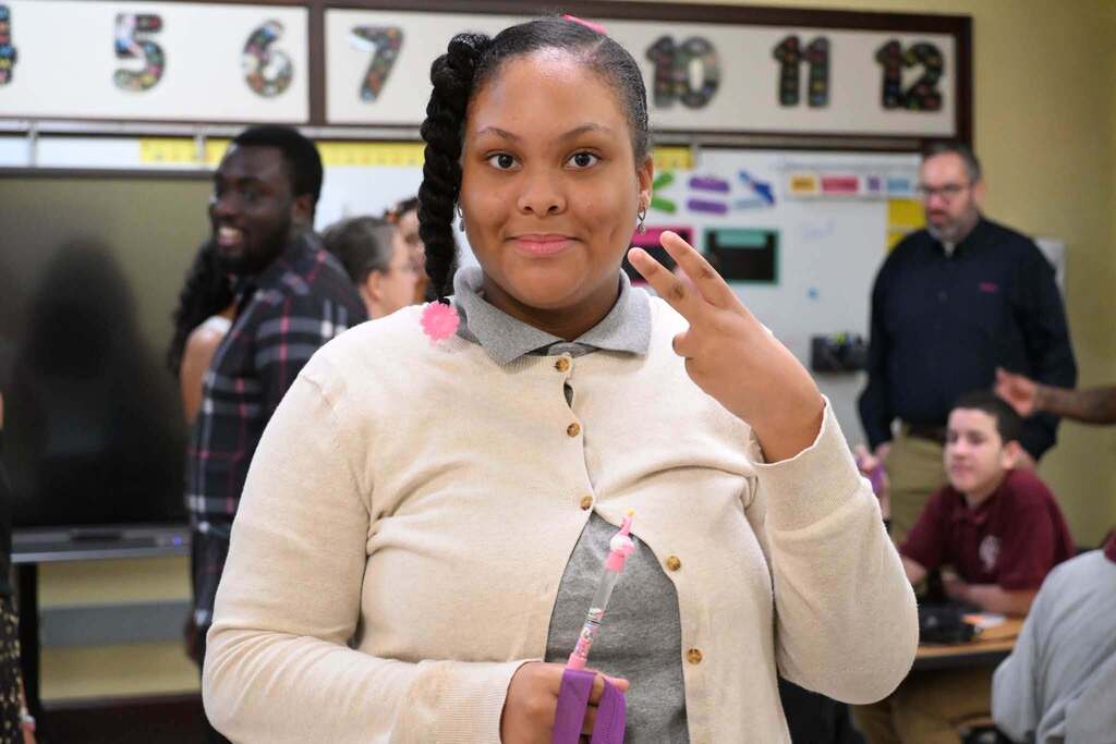 A student is smiling and holding up a peace sign with one hand while holding a pencil in another and standing in a school classroom. Other people can be seen nearby in the background.