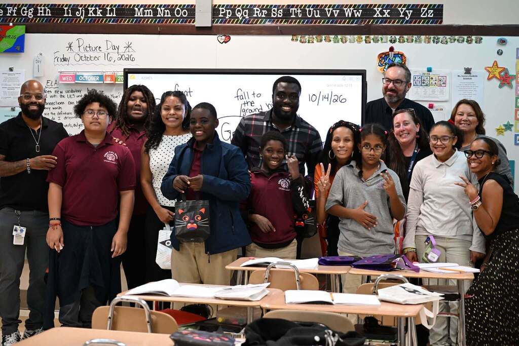 A group of district staff members, students, and community members are posing for a group photo in a school classroom, while smiling and holding colorful bags.