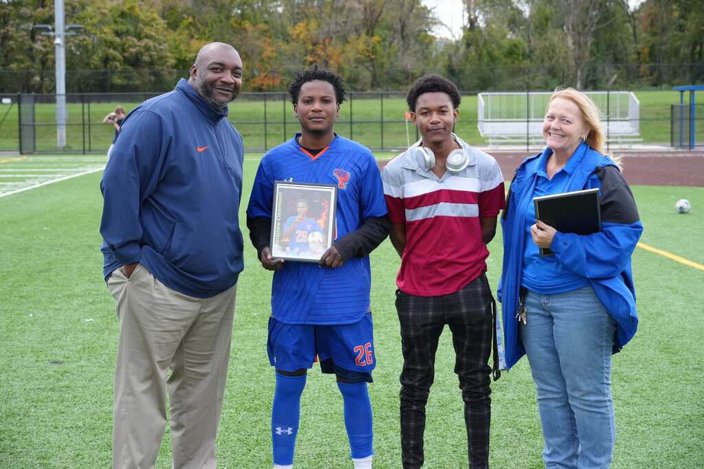 A William Penn boys' soccer senior is posing with a family member and district staff members, while holding a framed photo during Senior Night.