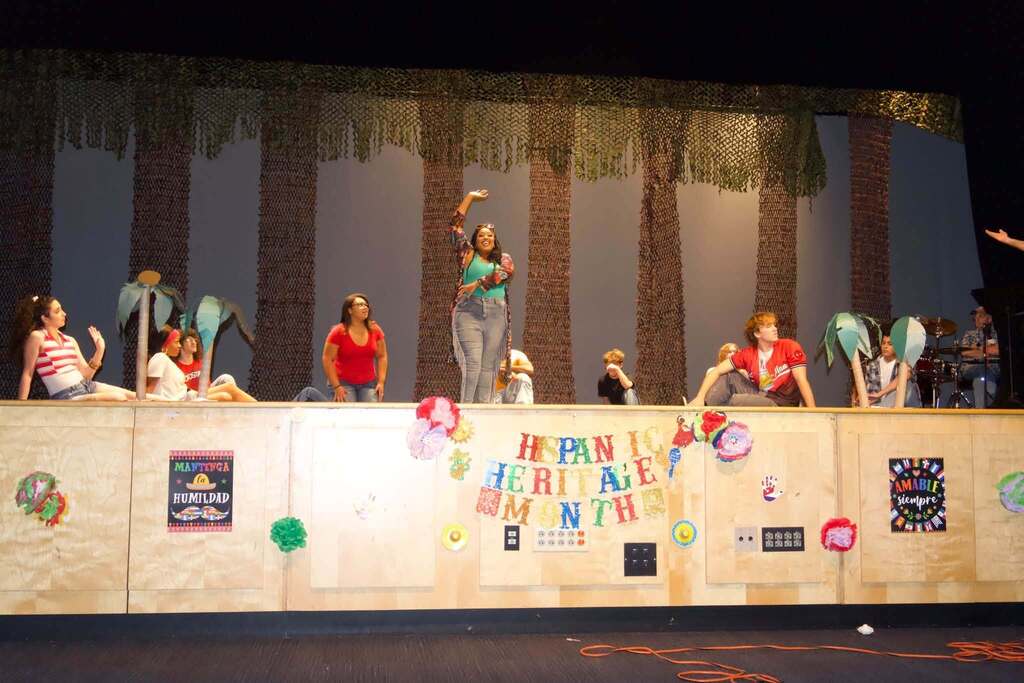 Students perform onstage during a Hispanic Heritage Month celebration, surrounded by colorful decorations and cultural displays.