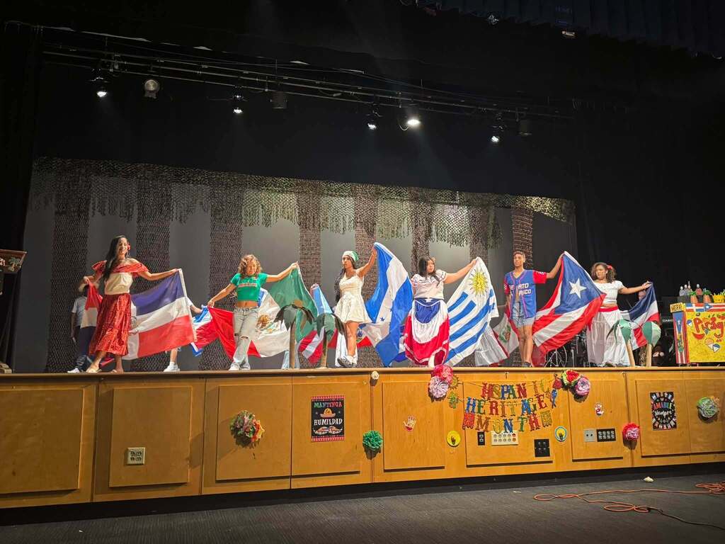 Students are celebrating Hispanic Heritage Month onstage, displaying flags representing various Latin American countries and Puerto Rico.