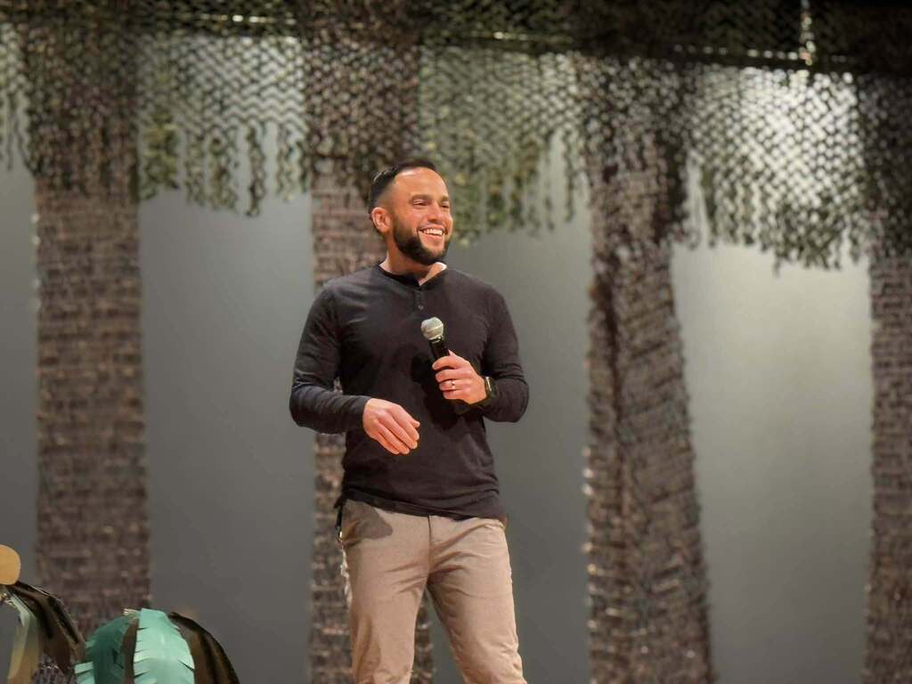 A guest speaker is speaking to an audience onstage during a Hispanic Heritage Month celebration decorated with colorful cultural displays.