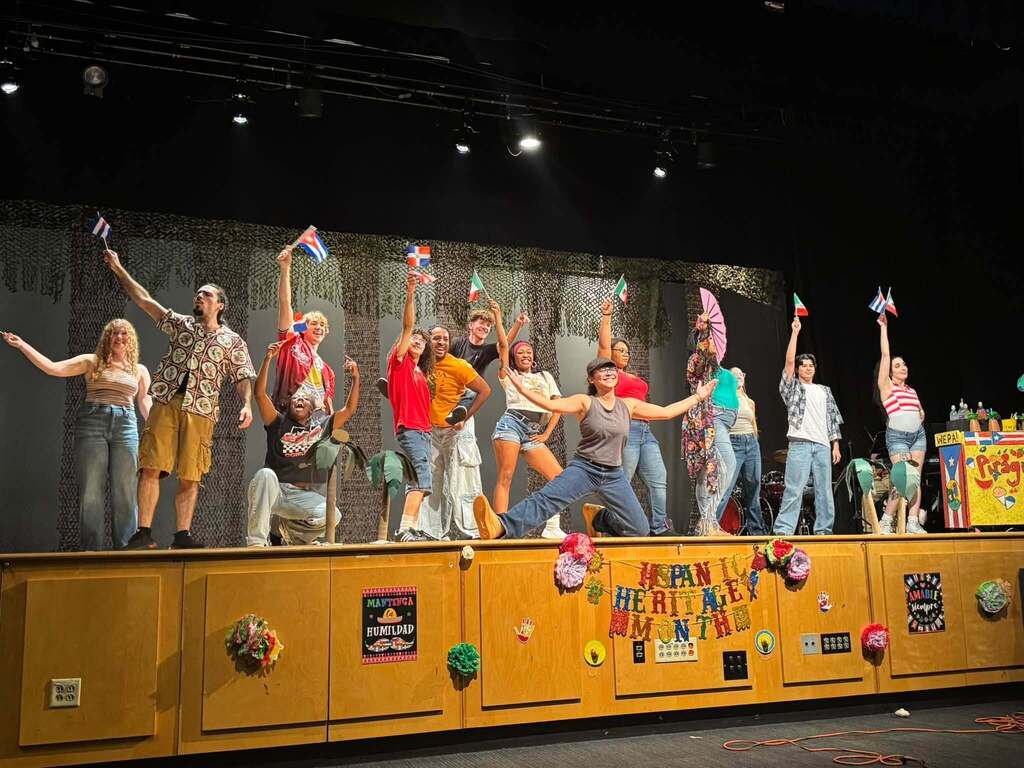 Students are performing onstage during a Hispanic Heritage Month celebration, surrounded by colorful decorations and cultural displays.