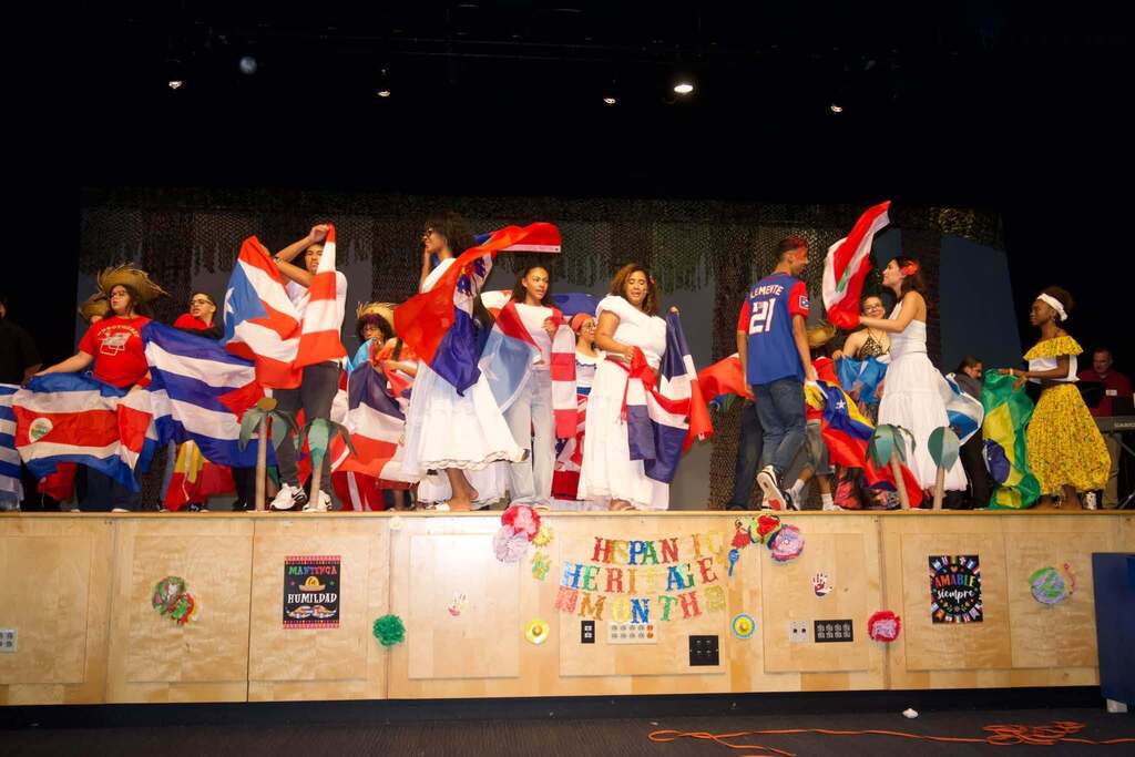 Students are celebrating Hispanic Heritage Month onstage, waving colorful flags and wearing traditional cultural attire.