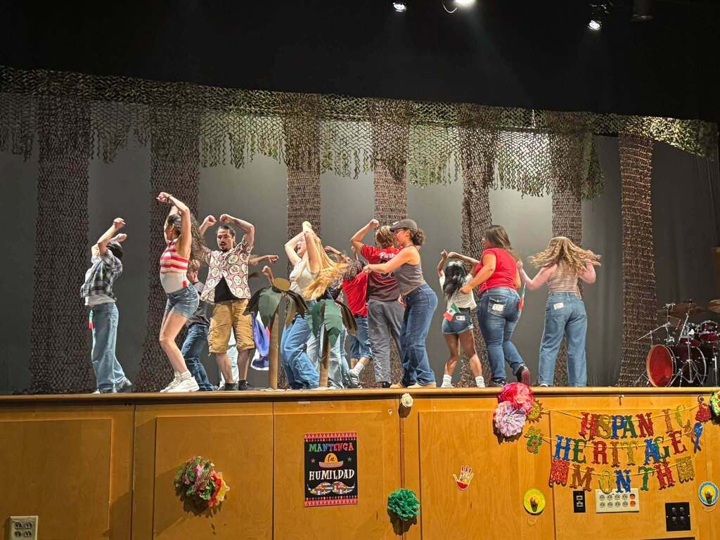 Students perform onstage during a Hispanic Heritage Month celebration, surrounded by colorful decorations and cultural displays.