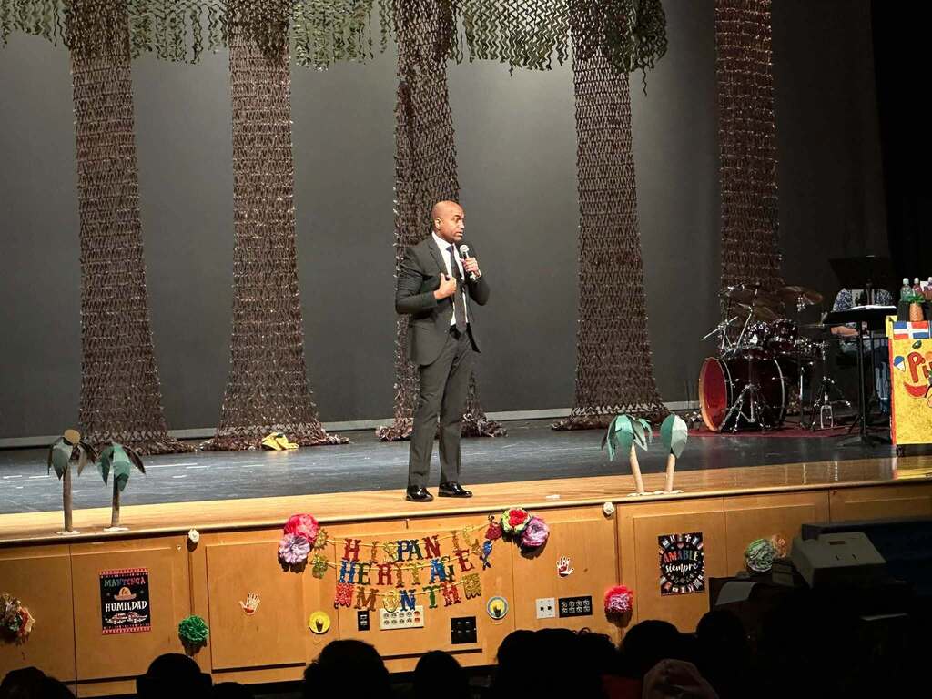 A guest speaker is speaking to an audience onstage during a Hispanic Heritage Month celebration decorated with colorful cultural displays.