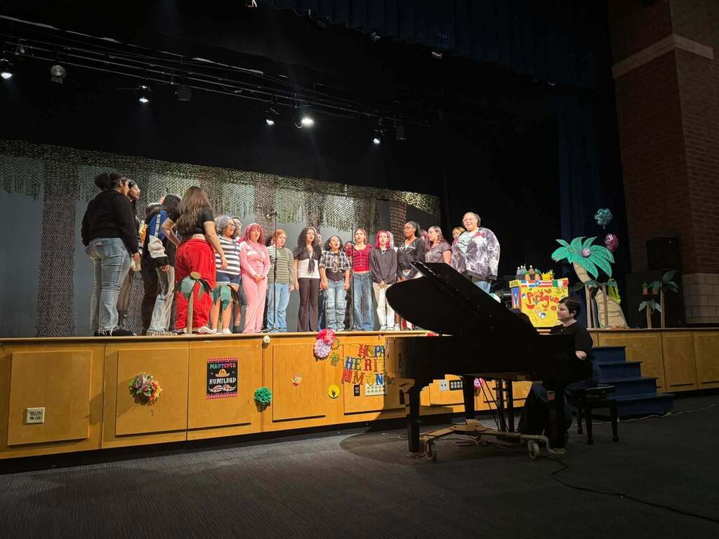 Students are performing onstage during a Hispanic Heritage Month celebration, accompanied by a pianist.