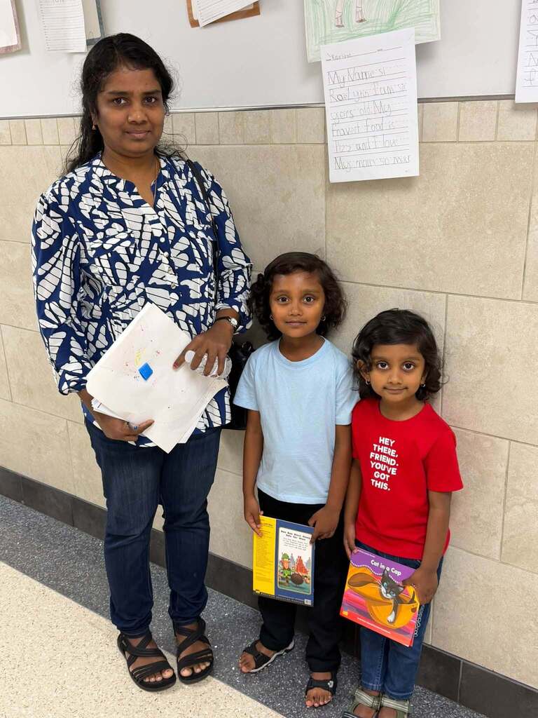 An adult woman and two young students are posing together. The two students are holding books in a school hallway decorated with student writing and artwork.