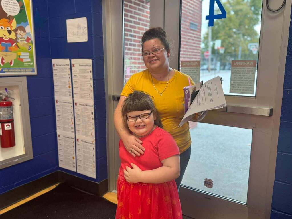 A smiling student in a pink dress is posing with a family member in a yellow shirt near the school entrance.