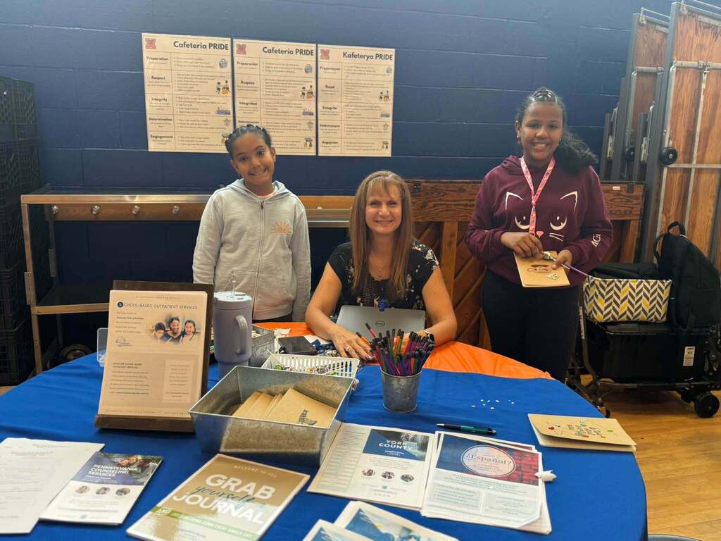 A staff member and two students are smiling behind a table displaying school-based support resources, informational brochures, and creative activities.