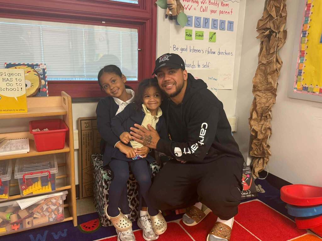 A smiling family member is posing with two students in a school classroom reading area.