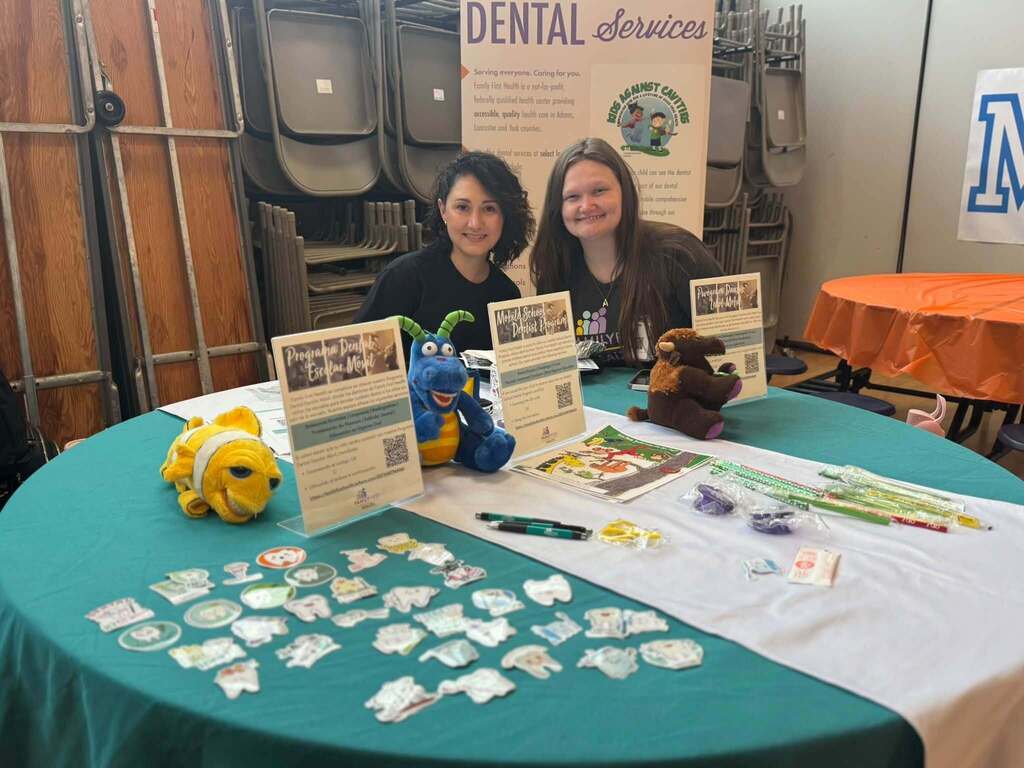 Two staff members are smiling behind a table displaying dental health resources, educational materials, and colorful plush characters.