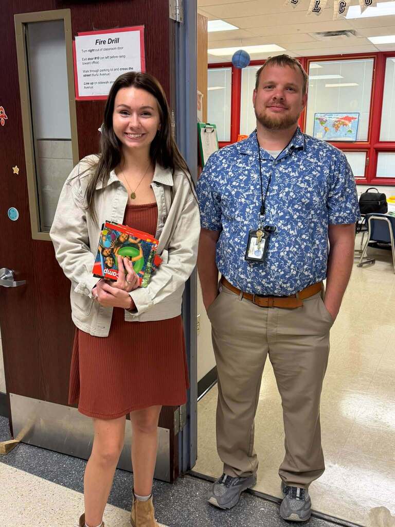 Two smiling school staff members are posing in a school classroom doorway, with one holding a stack of children’s books.