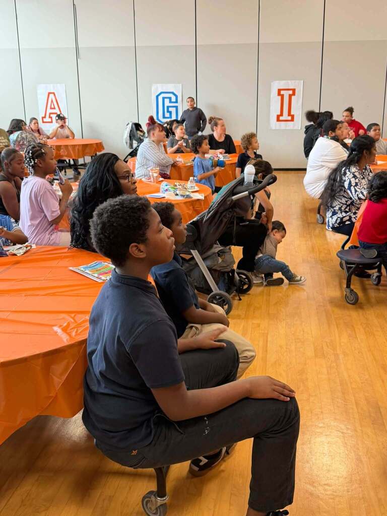 Students and families are gathering around orange-covered tables during a school community event, watching a presentation.