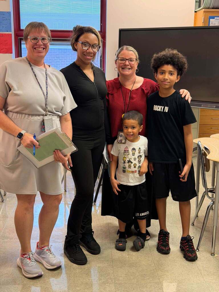 Two school staff members are posing with an adult woman and two young students in a school classroom.
