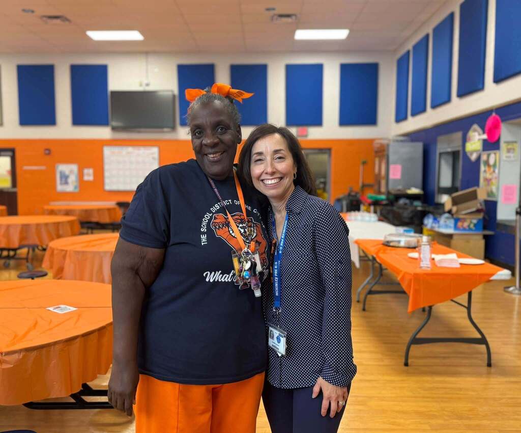 Two smiling school staff members are posing together in a school cafeteria decorated with orange and blue district colors.