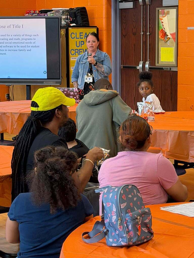 A district administrator is presenting information about the Title I program to families seated at orange-covered tables during a school event.