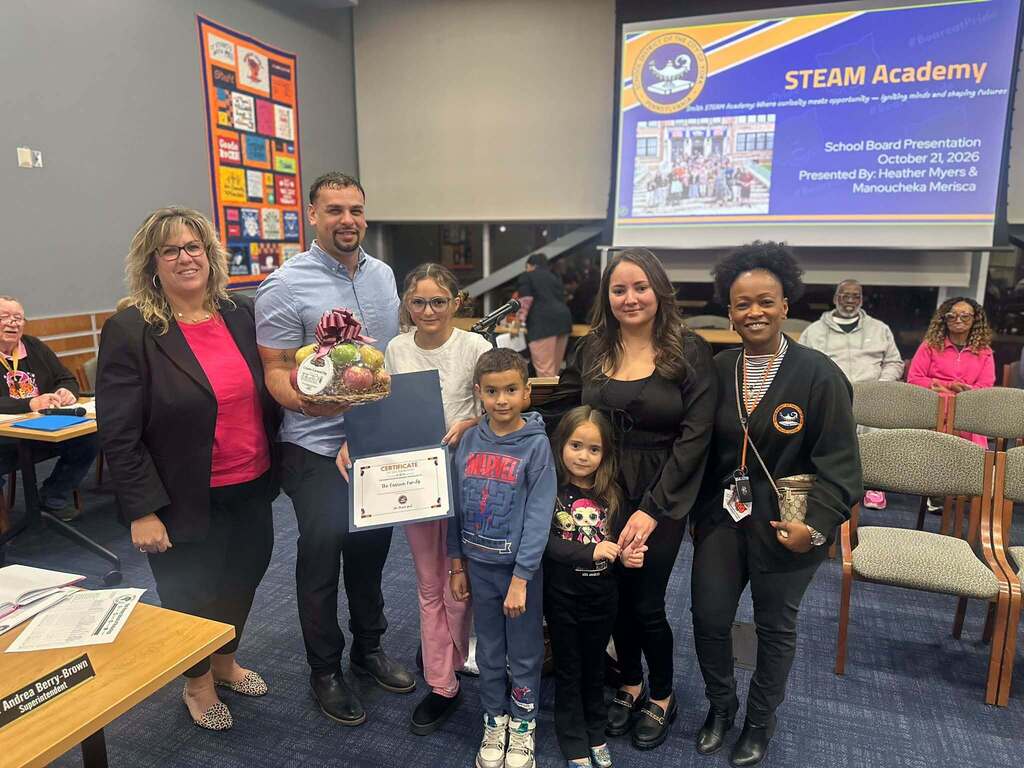 District staff and a district family are posing for a photo during a Smith STEAM Academy School Board presentation, celebrating family recognition with a certificate and gift basket.