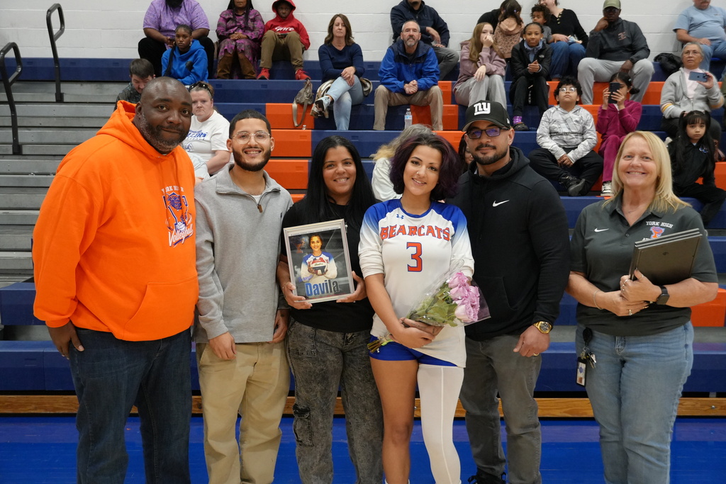 A Bearcats volleyball player is posing with her family and coaches during Senior Night, holding flowers and a framed photo, with spectators seated in the bleachers behind them.