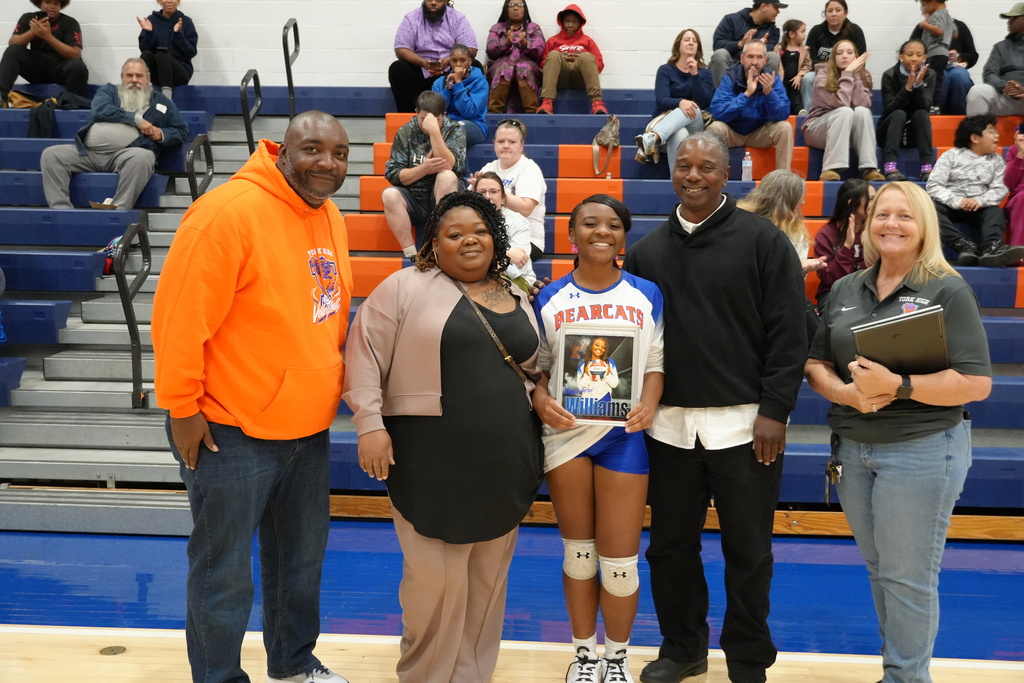 A Bearcats volleyball player is posing with her family and coaches on Senior Night, holding a framed photo of herself in uniform, as spectators watch from the bleachers.