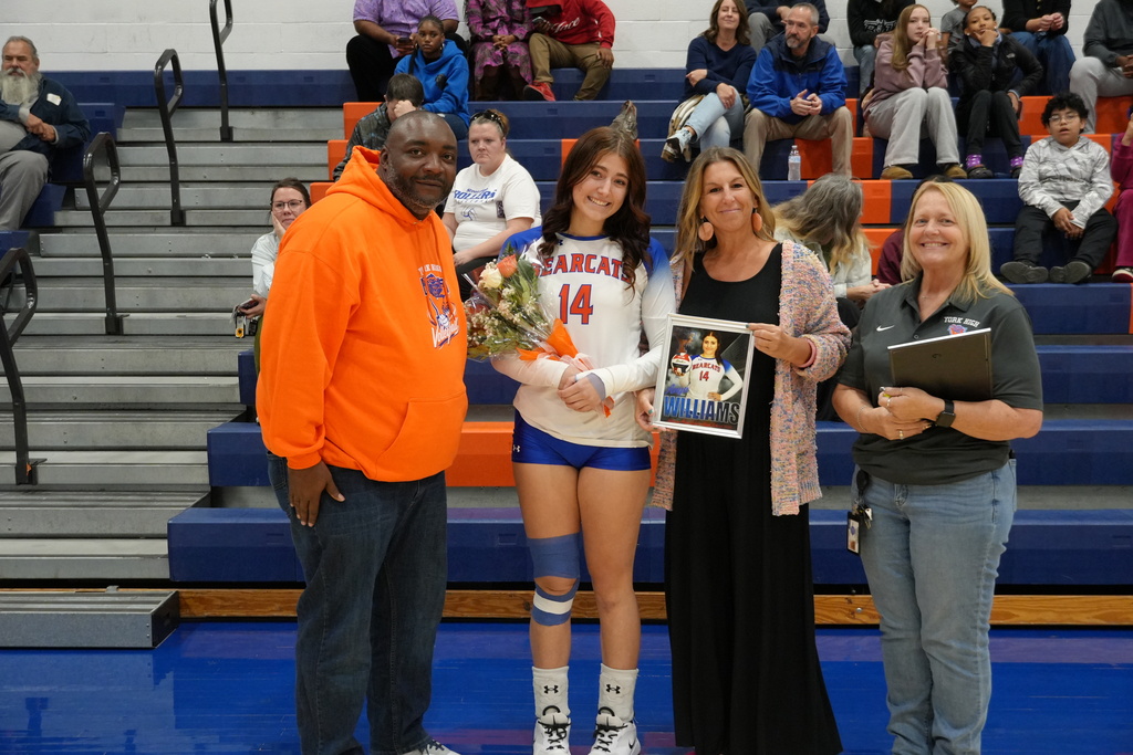 A Bearcats volleyball player is posing with her family and coaches on Senior Night, holding flowers alongside a framed photo of herself in uniform.