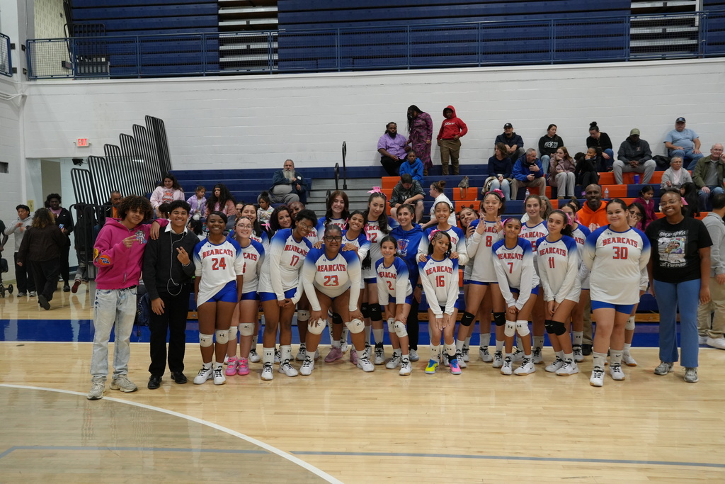 The Lady Bearcats Girls volleyball team is posing for a group photo with coaches and supporters during Senior Night in the school gymnasium.