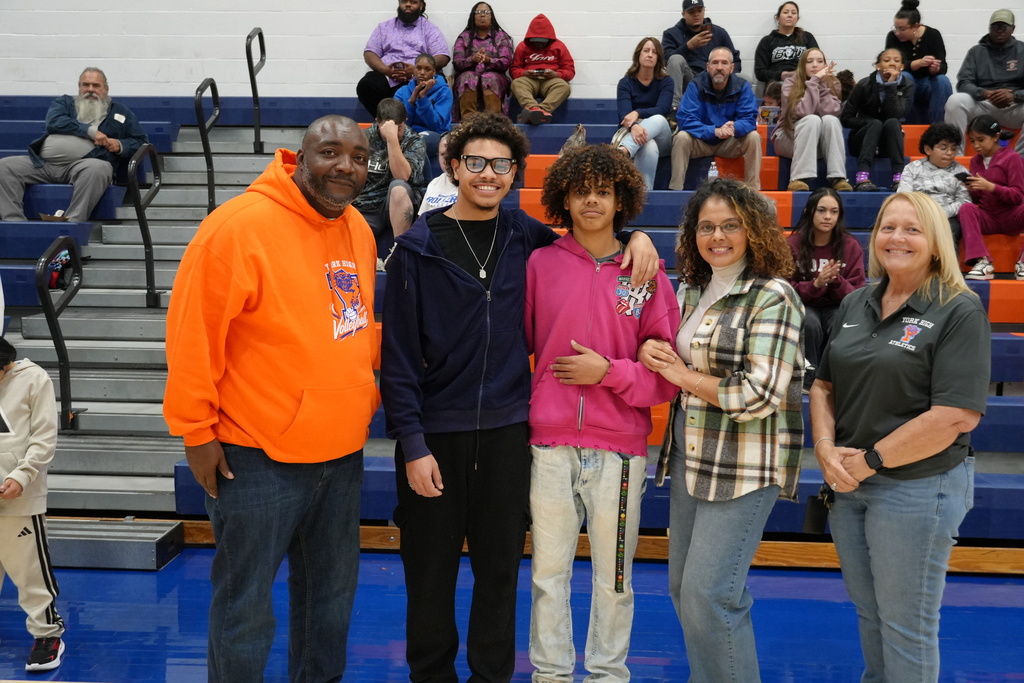 Two students are posing with their family and coaches during Bearcats volleyball Senior Night, smiling in front of the gymnasium bleachers.