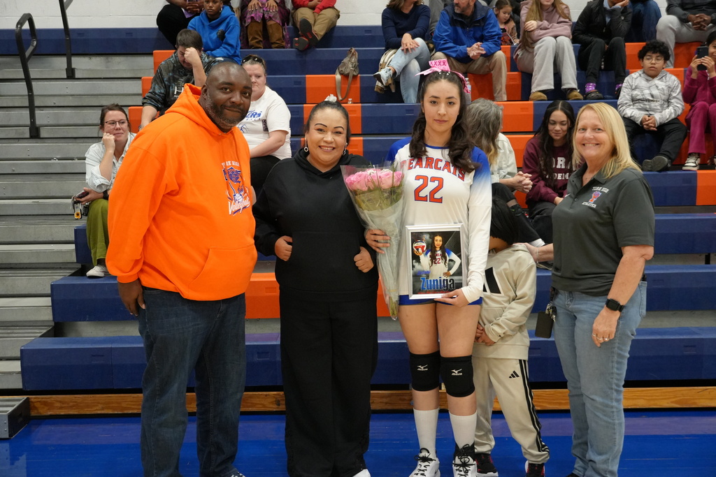 A Bearcats volleyball player is posing with her family and coaches on Senior Night, holding pink roses and a framed photo of herself in uniform.